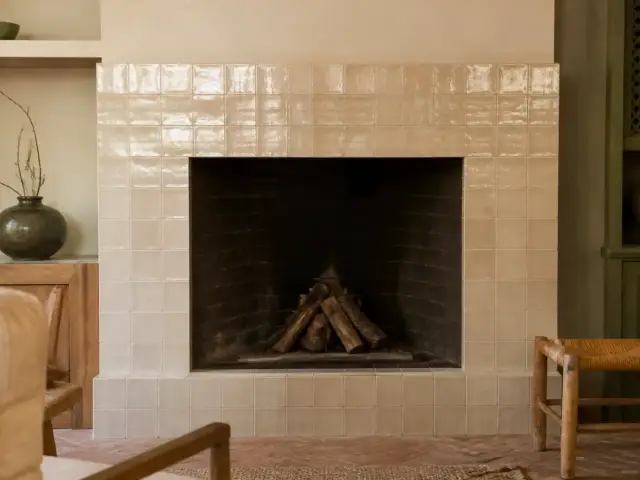 Manteau de cheminée habillé de carreaux de zellige beige brillant autour d’un foyer en briques sombres