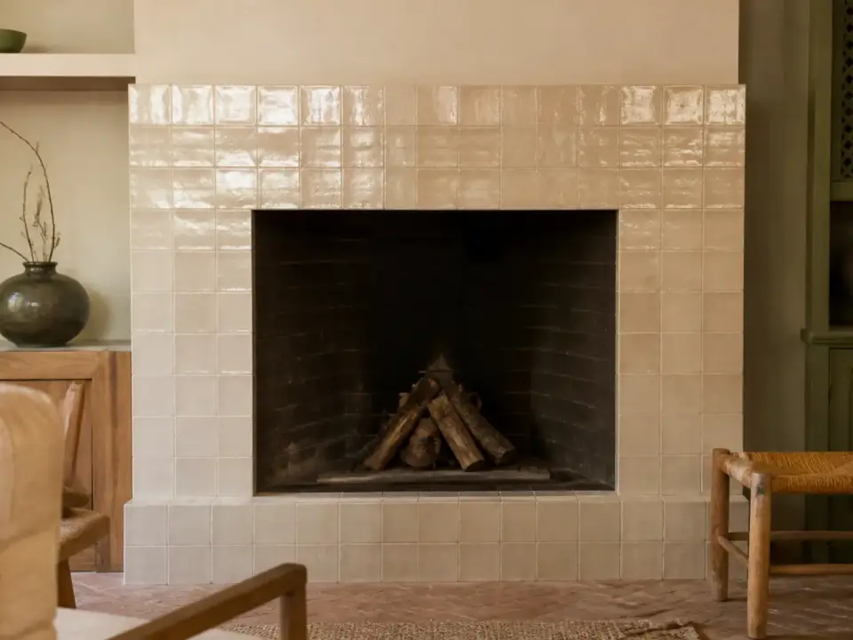 Manteau de cheminée habillé de carreaux de zellige beige brillant autour d’un foyer en briques sombres
