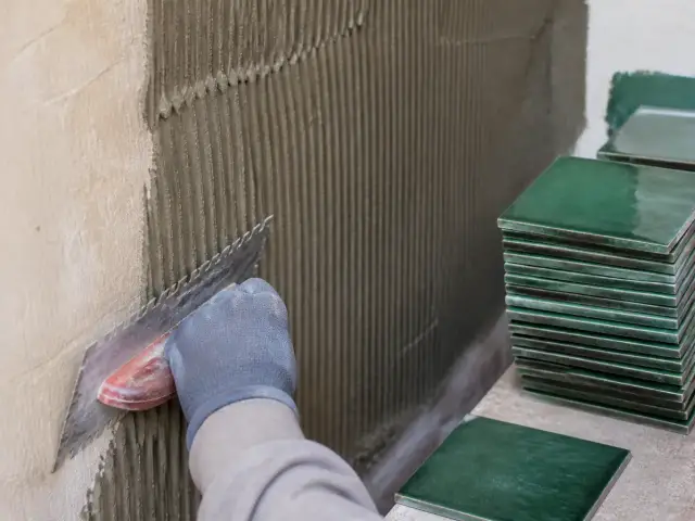 Peigne cranté étalant une colle grise en sillons sur un mur, carreaux de zellige vert empilés à côté